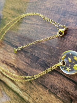 Collier "Echos de l'océan" transparent composé d'une chaîne et d'un pendentif en acier inoxydable de couleur dorée rond et plat, transparent, dans lequel sont inclus de petits éléments marins.