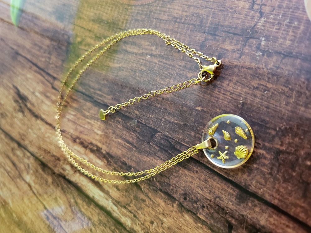 Collier "Echos de l'océan" transparent composé d'une chaîne et d'un pendentif en acier inoxydable de couleur dorée rond et plat, transparent, dans lequel sont inclus de petits éléments marins.