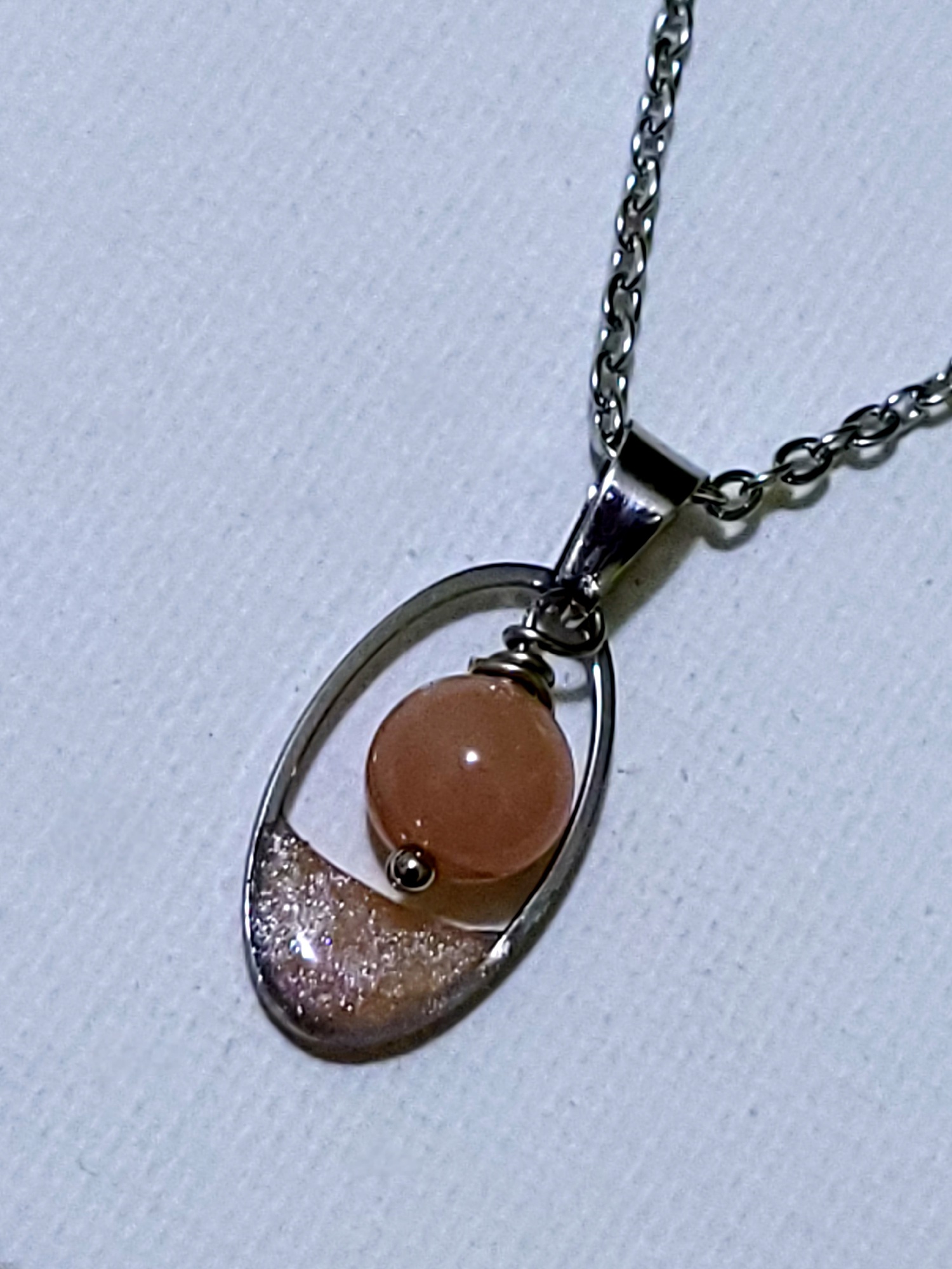 Collier Goutte Pierre de Soleil de Perle et Coquette : pendentif ovale en acier avec perle de pierre de soleil naturelle et résine corail pailletée.